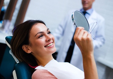 Woman smiling in dental mirror after smile makeover in Garland