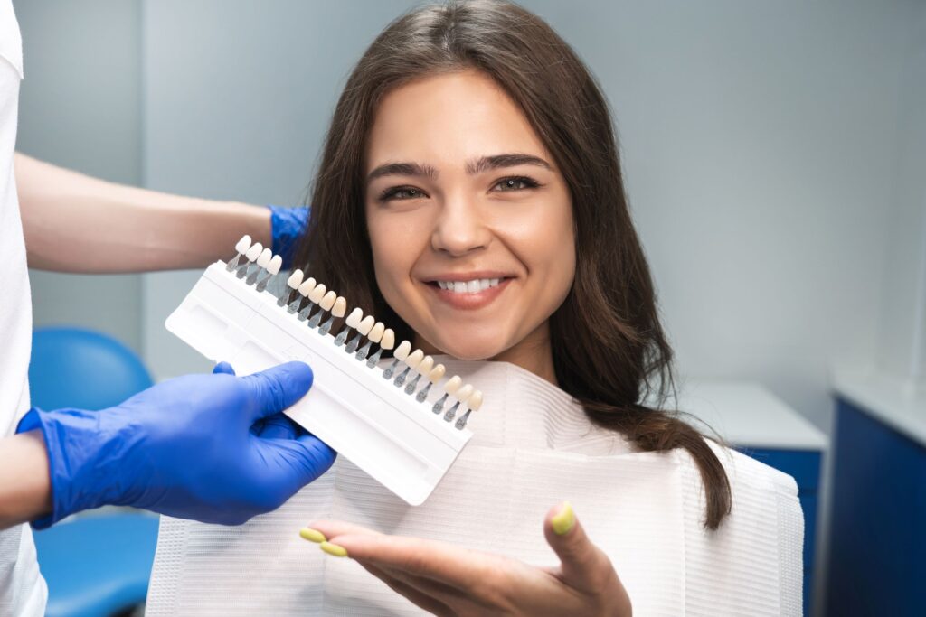 Woman with brown hair and yellow nails gesturing to shade guide held to teeth